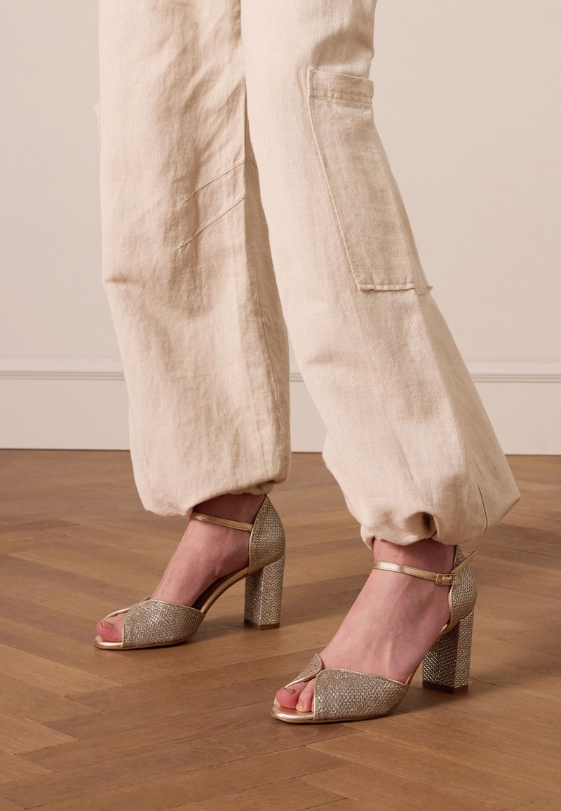 Gestreepte hoge hakken in glanzend gouden gaas met een chunky blokhak en open teen ontwerp, gecombineerd met lichtbeige, losse broek.