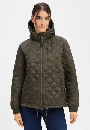 Woman wearing a green quilted hooded jacket with front zipper and pockets, paired with dark blue jeans, against a plain background.