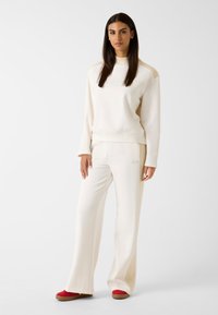 Kremowy zestaw wypoczynkowy składający się z długiego rękawa bluzki z prążkowanymi akcentami i dopasowanych spodni. Czerwone buty wsuwane dopełniają stylizację.