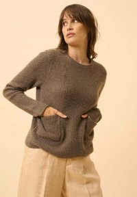 Brauner gerippter Strickpullover mit langen Ärmeln und zwei Fronttaschen, kombiniert mit hellbeigen, weit geschnittenen Hosen vor einem neutralen Hintergrund.