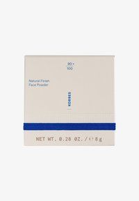 Boîte rectangulaire blanche avec des accents bleus. Le texte indique "Poudre de visage à finition naturelle", et les détails du produit sont imprimés en bleu et marron.