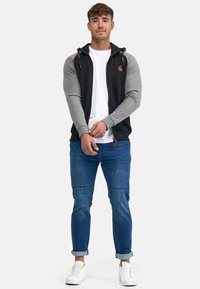 Chaqueta con cremallera para hombre de cuerpo negro y mangas grises, que cuenta con cuerda ajustable y un acento de logo. Combinada con jeans azules y zapatillas blancas.