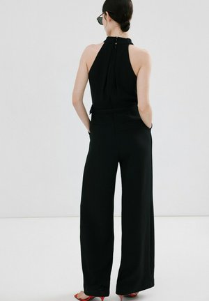 Vrouw met donker haar in een knot, gekleed in een zwarte mouwloze halterjumpsuit en rode open-toe hakken, staand met handen in de zakken en met de rug naar de kijker.