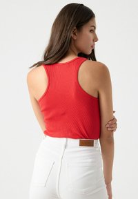 Top de tirantes rojo con canalé y amplios huecos para los brazos, combinado con unos jeans blancos de tiro alto que tienen una etiqueta de cuero marrón con la inscripción "INSIDE".