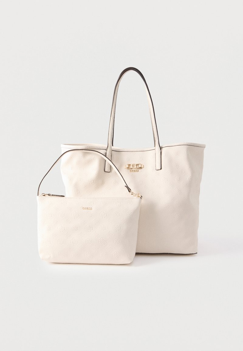 Δύο τσάντες Guess σε κρεμ απόχρωση: μια μεγάλη τσάντα ώμου με διπλές λαβές και μια μικρότερη τσάντα με φερμουάρ και ένα λουράκι, και οι δύο με ανάγλυφα λογότυπα.