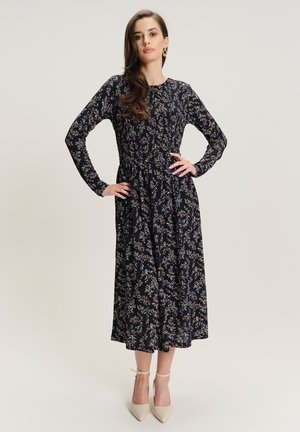 Vestido floral azul marino con mangas largas, cintura fruncida y falda amplia. Presenta un delicado patrón de flores en varios colores. Combinado con tacones beige.