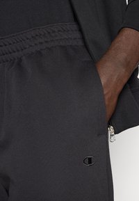 Μαύρο αθλητικό παντελόνι από υφασμάτινη υφή, με ελαστική μέση και φερμουάρ σε τσέπη, διακοσμημένο με μικρό λογότυπο.