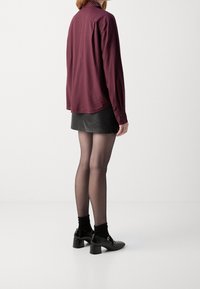 Camisa de botón de color burdeos con estampado y mangas largas, combinada con una falda de cuero negra, medias transparentes y zapatos negros de tacón.