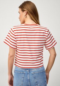 Červený a bílý pruhovaný crop top, krátké rukávy, bavlněný materiál, kulatý výstřih, hladká textilie, nošený s modrými džíny.