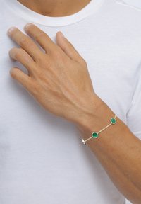 Hand mit einem silbernen Kettenarmband, das zwei grüne runde Steine zeigt, die auf einem weißen Hemd in Brusthöhe ruhen.