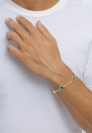 Hand mit einem silbernen Kettenarmband, das zwei grüne runde Steine zeigt, die auf einem weißen Hemd in Brusthöhe ruhen.