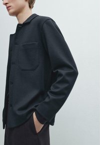 Young person wearing a black buttoned jacket with a chest pocket, standing with one hand in dark pants pocket against a plain background.