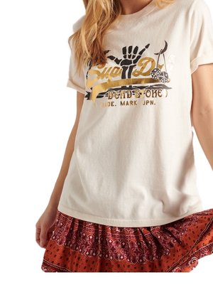 Frau trägt ein weißes Grafik-T-Shirt mit schwarzem und goldenem Druck und einen roten gemusterten Rock, hält den Rocksaum.