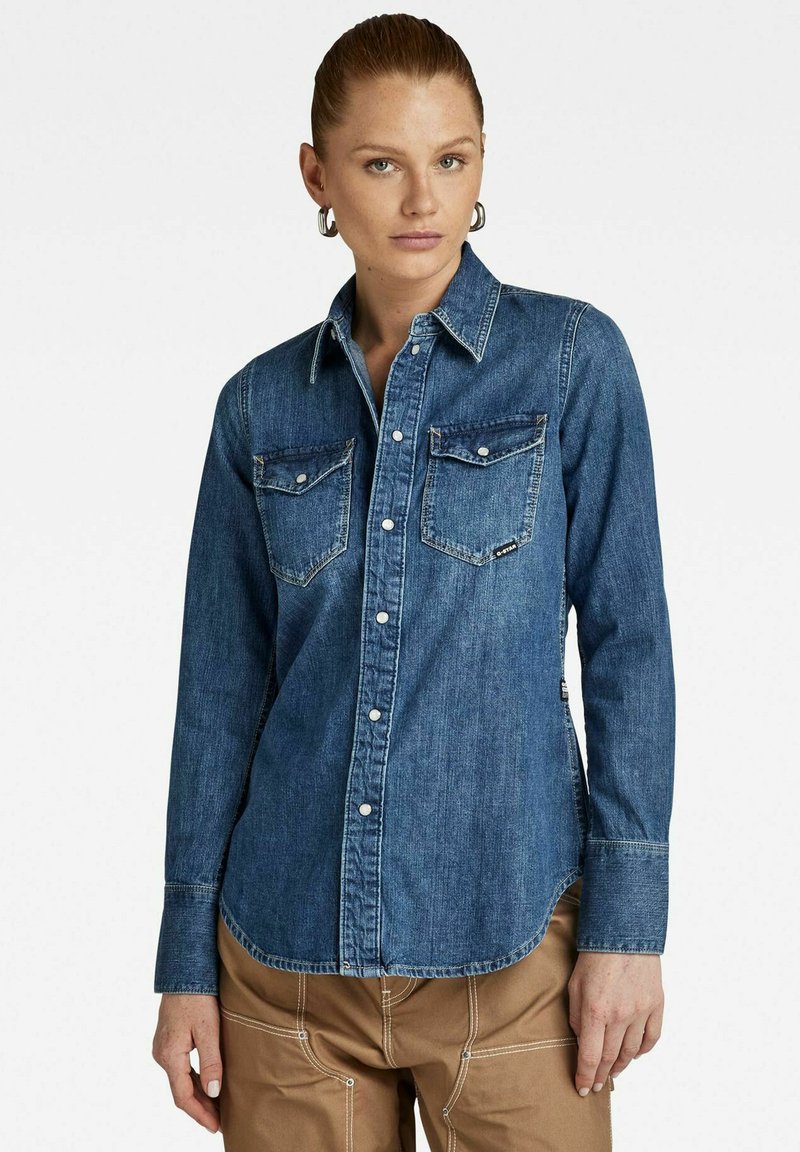 Camicia in denim a maniche lunghe di colore blu scuro, con colletto a punta, due tasche sul petto e bottoni argentati. Tessuto morbido con dettagli di cucitura discreti.