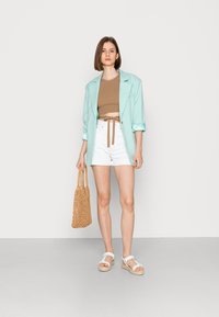 Blazer azul claro, top cropped beige com um laço, calções brancos e sandálias brancas. Modelo segurando uma bolsa tecida beige. Fundo suave.