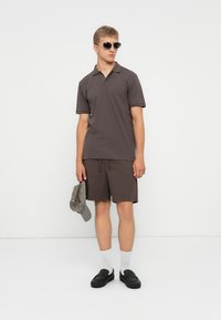 T-shirt polo marron à manches courtes et col, associé à un short assorti. Le modèle tient une casquette grise, porte des lunettes de soleil et des chaussures noires.