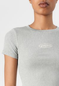 Maglietta grigia a costine con scollatura rotonda e maniche corte; presenta un logo "Guess Jeans" beige chiaro sul petto.