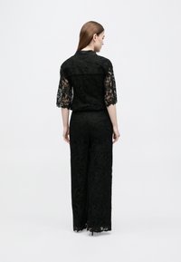 Γυναίκα όρθια, φοράει μαύρο floral δαντελένιο παντελόνι με φαρδύ πόδι και ταίριαστο δαντελένιο τοπ με μανίκια μήκους μέχρι τον αγκώνα, γυρισμένη με την πλάτη προς λευκό φόντο.