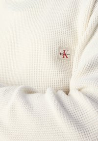 Close-up van witte wafelstructuurstof met een klein Calvin Klein logo-op-patch in rode en zwarte stiksels.