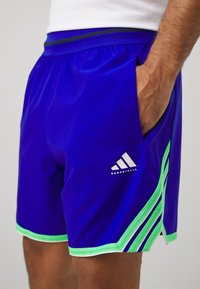 Pantalones cortos deportivos azules con acentos verdes, que cuentan con una cintura elástica, bolsillos laterales y un logo de Adidas con el texto "BASKETBALL".