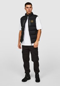 Zwarte gewatteerde vest met een hoge kraag, rits aan de voorkant en logopatch. Draag het over een wit T-shirt en combineer het met zwarte cargobroek en sneakers.