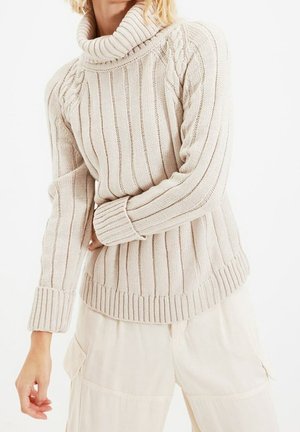 Chunky knit beige sweater with a high collar, ribbed texture, and cable details on shoulders, paired with light-colored pants.