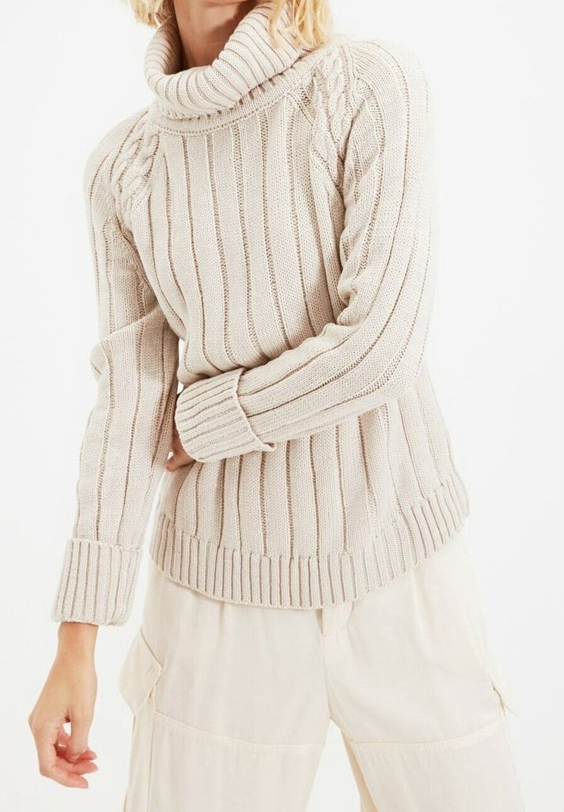 Chunky knit beige sweater with a high collar, ribbed texture, and cable details on shoulders, paired with light-colored pants.