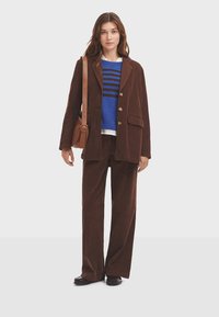 Brauner Cord-Blazer und passende weit geschnittene Hose, darüber ein blauer gestreifter Pullover getragen. Dunkle Schuhe und eine beige Umhängetasche vervollständigen den Look.