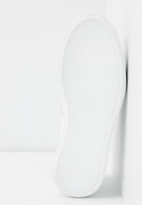 Bílá podrážka tenisky s jemnými horizontálními drážkami, umístěná svisle na bílém povrchu.