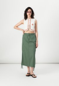 Geknipt wit topje met oranje stiksel en zonnenembleem, gecombineerd met een groene enkellange rok met zijzakken en details met trekkoord.