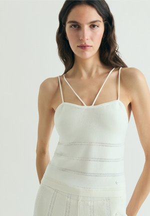 Mujer con cabello largo y oscuro, vestida con una blusa blanca sin mangas de tirantes finos y pantalones blancos a juego, frente a un fondo liso.