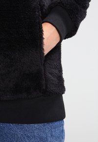 Käsi mustan pörröisen takin taskussa, takki on yhdistetty sinisiin farkkuihin, ja taustana on vaalea yksivärinen pinta.