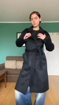 Ung person som justerer kragen på en lang svart kåpe med belte, stående i stuen med grønn vegg, brun sofa og tregulv.