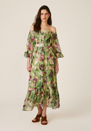 Mujer con vestido maxi verde con flores y escote de hombros descubiertos, con cinturón en la cintura y tacones abiertos marrones, de pie frente a un fondo liso.