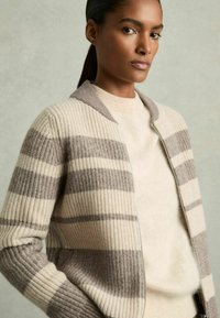 Randig cardigan i krämfärg och taupe, ribbad textur, med dragkedja och bred krage. Bärs över en ljus beige tröja.