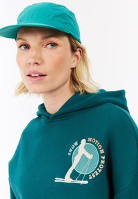 Tyrkysová mikina s kapucňou so sústredným logom znázorňujúcim lyžiara a textom, v kombinácii so svetlomodrou čiapkou. Mäkkučký materiál, voľný strih, tonálna niť.