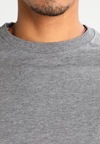 Sudadera gris con cuello redondo acanalado, tejido texturizado y ligeras variaciones en el tono, mostrando el cuello y la parte superior del pecho.