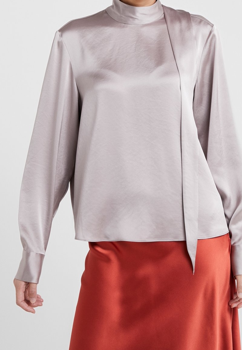 Femme portant un chemisier en satin gris clair à manches longues avec un col montant et un détail noué, associé à une jupe en satin rouge.