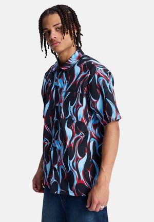 Młody mężczyzna z warkoczami, ubrany w koszulę z krótkim rękawem w czerwono-niebiesko-czarny wzór płomieni oraz ciemnoniebieskie dżinsy.