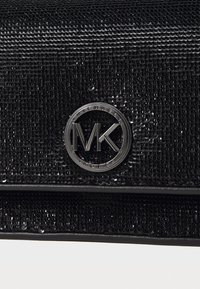 Sac à main noir texturé avec une finition en sequins brillants et un logo circulaire en argent présentant "MK" et "Michael Kors" au centre.
