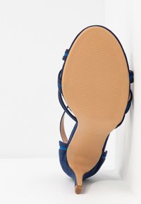 Semelle d'une sandale à talon aiguilles bleu marine avec une semelle extérieure texturée couleur fauve et un talon fin, inclinée contre un fond blanc en coin.