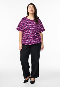 Kurzarmoberteil mit einem rosa und lila geometrischen Muster auf dunklem Hintergrund, kombiniert mit schwarzen weiten Hosen und schwarzen Schuhen mit hohen Absätzen.