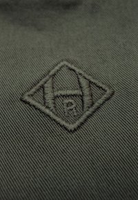 Dunkelgrüner Stoff mit einer strukturierten Oberfläche, auf der ein gesticktes Diamantlogo zu sehen ist, das winkelige Formen und ausgeprägte Buchstabendetails integriert.