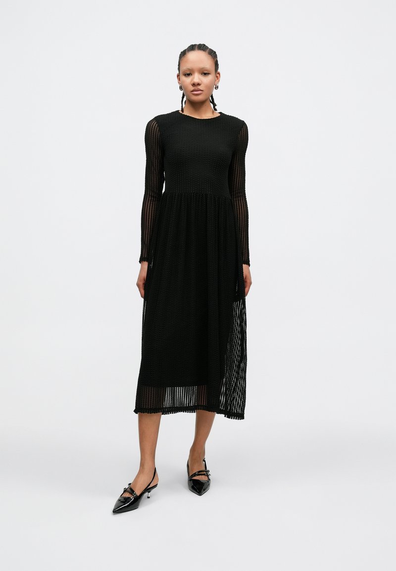 Black midi dress featuring a textured, ribbed design with long sleeves and a flared skirt. Paired with pointed black shoes.