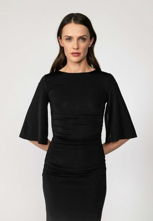 Kobieta o długich brązowych włosach, ubrana w dopasowaną czarną sukienkę z rozkloszowanymi rękawami do pół łokcia, stojąca na tle jasnego, jednolitego tła.