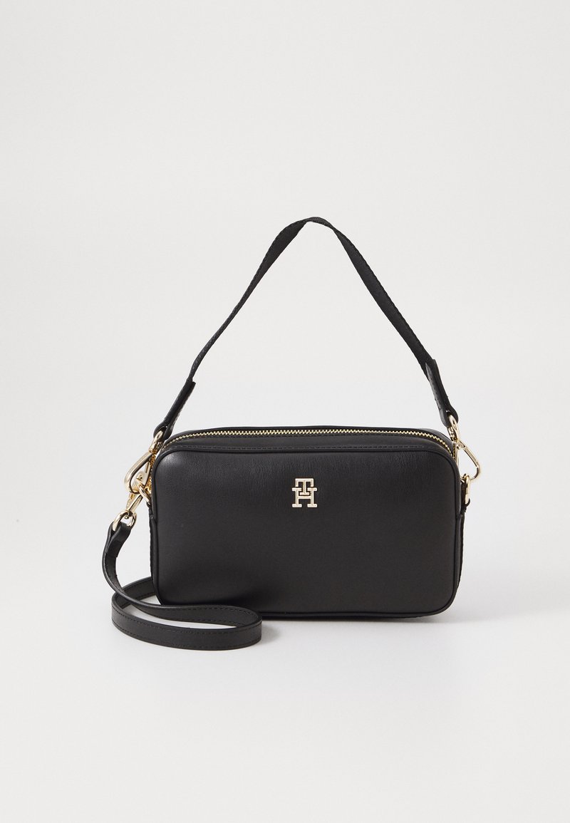 Sac à main en cuir noir avec quincaillerie dorée, fermeture éclair et bandoulière amovible. Présente un logo bien en vue à l'avant. Forme rectangulaire compacte.