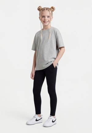 Fiatal lány dupla fonott kontyokkal, szürke túlméretezett pólót, fekete leggingset és fehér Nike tornacipőt viselve, egyik kezét a zsebében tartva áll.