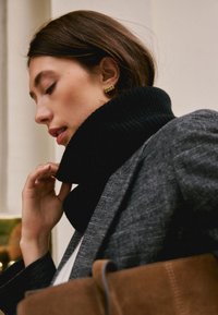 Crni rebrasti pulover s visokim ovratnikom, sivi karirani sako, zlatna naušnica u obliku obruča i smeđa antilop torba s kožnim remenom, pogled sa strane.