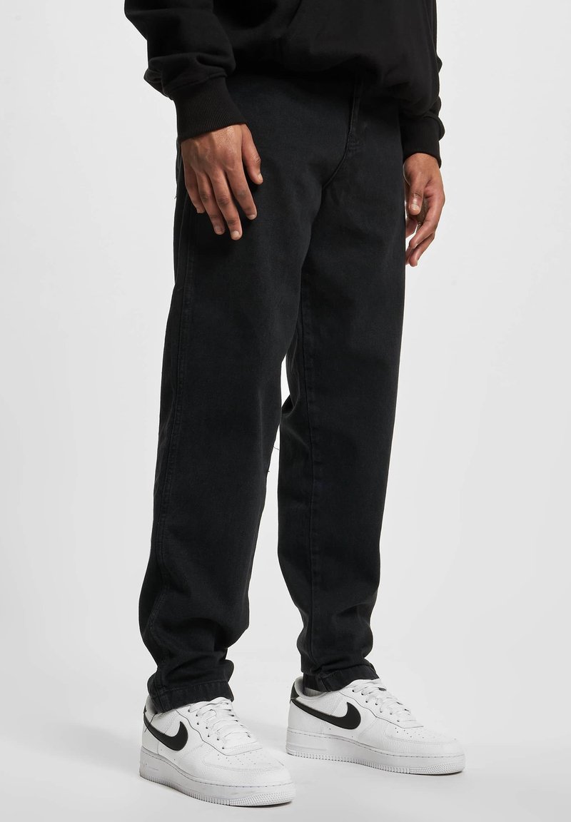 Personne portant un pantalon noir et des baskets blanches Nike avec un swoosh noir, debout devant un fond clair uni.