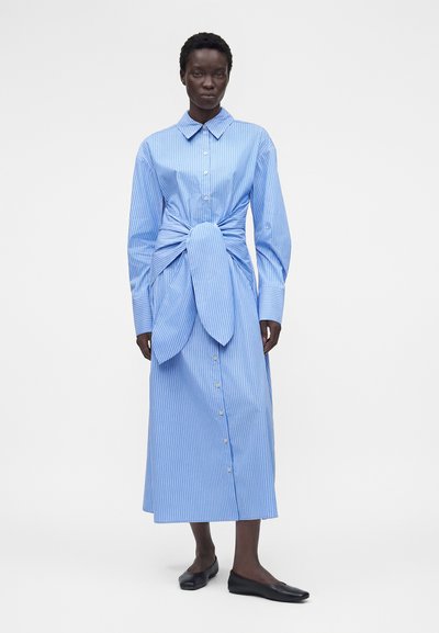 Γυναίκα που φοράει μακρύ μπλε και άσπρο ριγέ φόρεμα με κουμπιά και μεγάλο δεμένο κόμπο στη μέση και μαύρα επίπεδα παπούτσια.